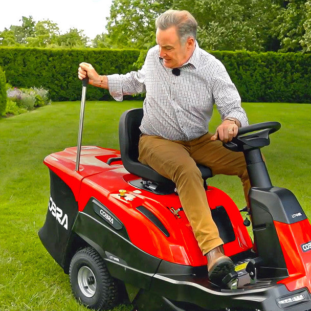 Man operating a red Cobra riding lawn mower on a green lawn with trees in the background.