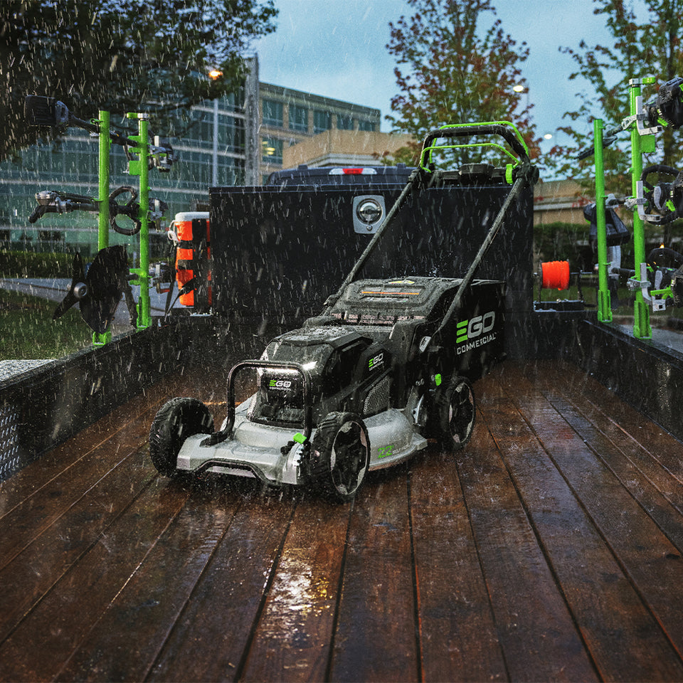 A wet EGO LMX5300SP cordless lawnmower shown on a wet wooden dock or platform outdoors in the rain, with a black container, green posts, trees, and a building in the background.