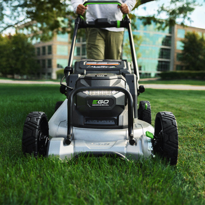 A person pushing a EGO LMX5300SP black and silver lawnmower with green accents on a green lawn, with a modern glass building in the background.